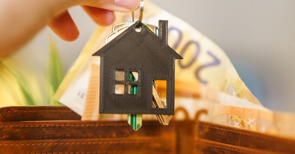 Hinnoittelu A hand holds a house-shaped keychain above an open wallet with Euro banknotes.
